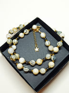 Collier Rivage en perles de coquillage blanches et grises, avec perles hématite noires et finitions dorées, présenté dans une boîte cadeau noire.