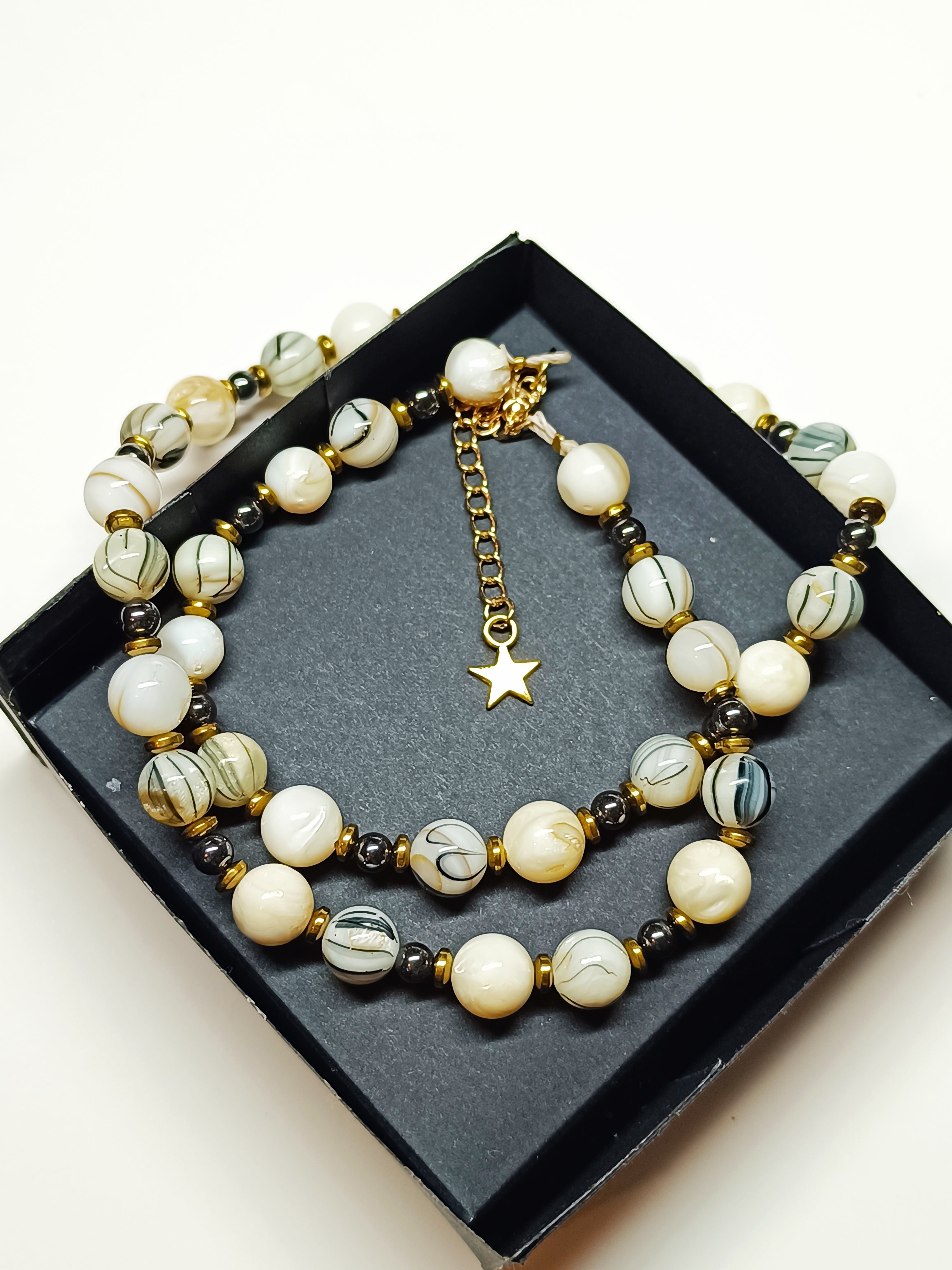 Collier Rivage en perles de coquillage blanches et grises, avec perles hématite noires et finitions dorées, présenté dans une boîte cadeau noire.