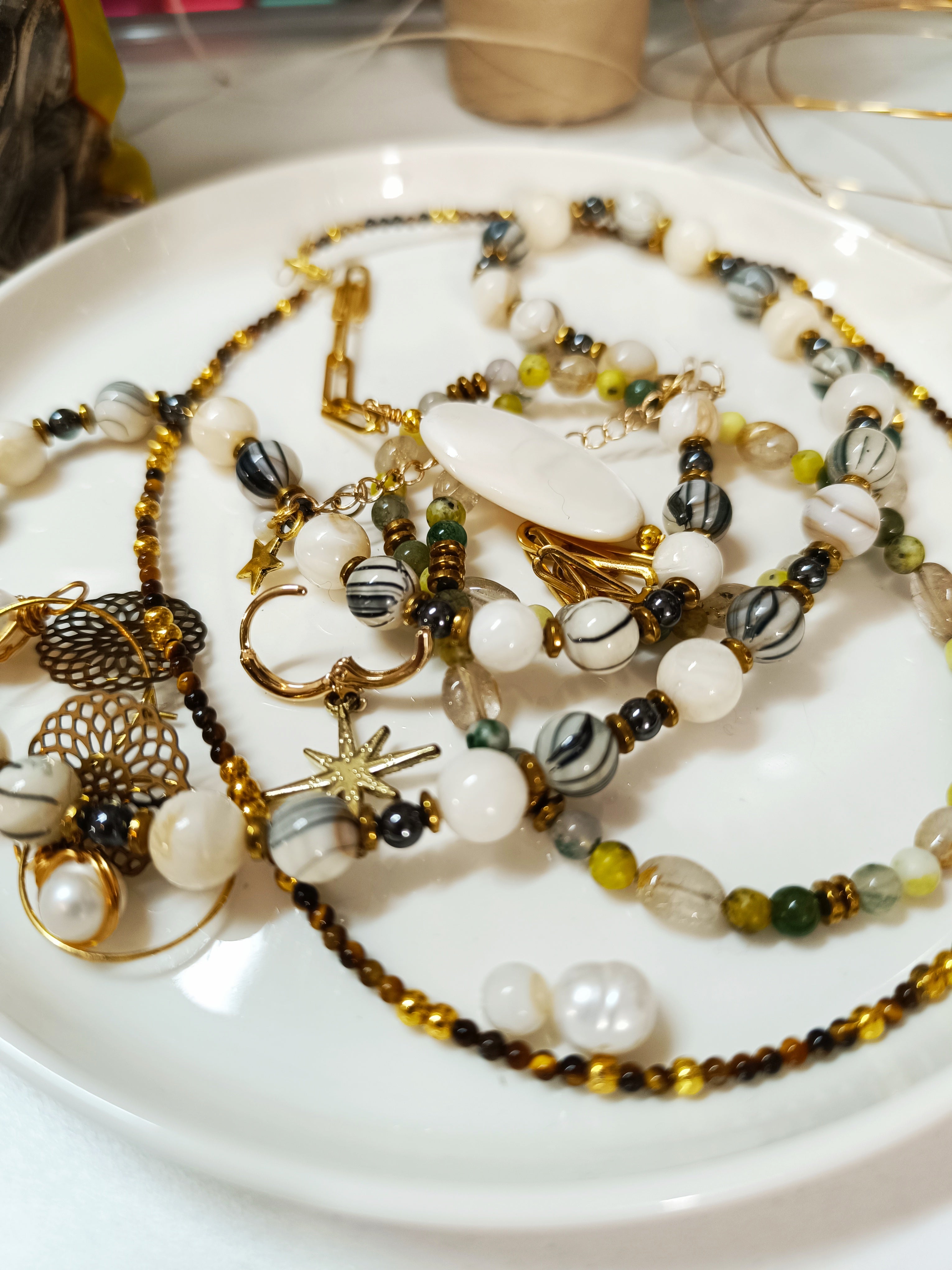 Assortiment de bijoux artisanaux dorés : colliers en perles de coquillage Rivage, bracelets Agate Mousse avec pendentif libellule, et autres créations en pierres naturelles et acier inoxydable.

