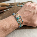 Bracelet multi-tours Luz pour homme en jaspe turquoise et perles dorées, porté au poignet sur un établi d'artisan avec outils de bijouterie en arrière-plan.