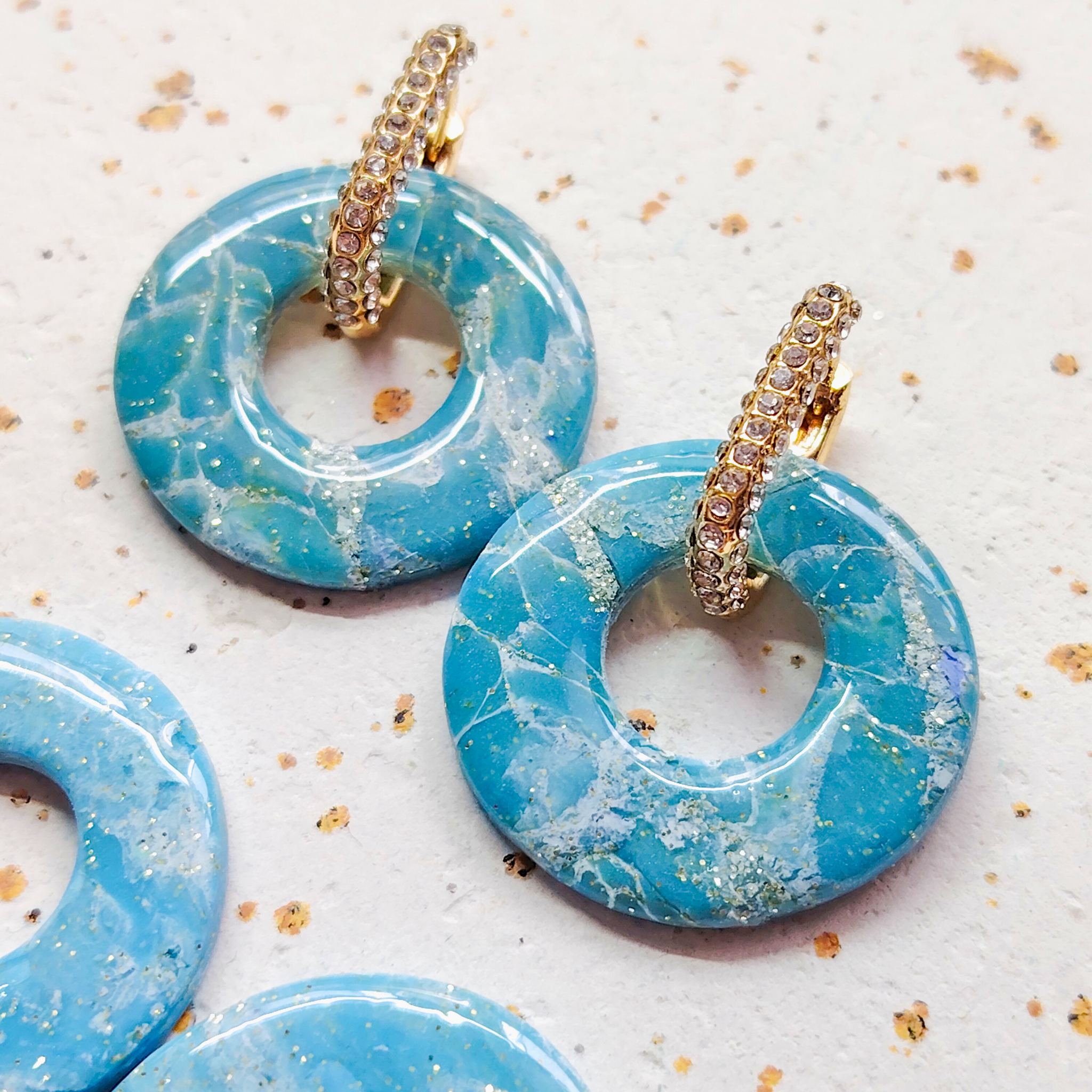 Gros plan lumineux sur une paire complète de boucles d'oreilles modulables Agate de la marque Sanaca, associant de larges pampilles effet pierre marbrée bleu et des créoles dorées ornées de strass.