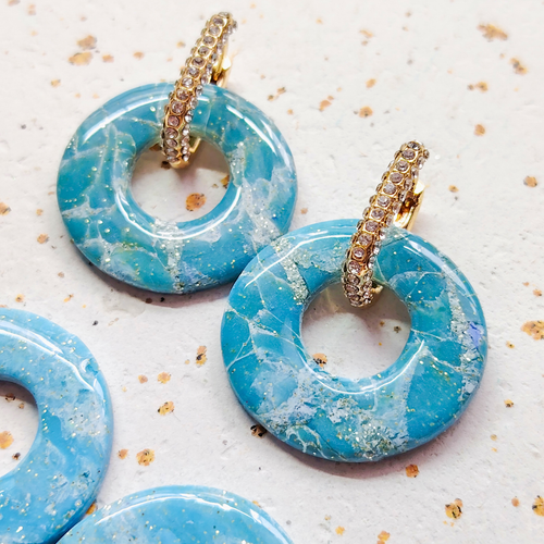 Gros plan lumineux sur une paire complète de boucles d'oreilles modulables Agate de la marque Sanaca, associant de larges pampilles effet pierre marbrée bleu et des créoles dorées ornées de strass.