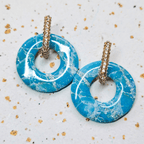 Paire de boucles d'oreilles créoles strass dorées avec leurs pampilles interchangeables Agate bleues et blanches aux reflets marbrés et pailletés d'or, posée sur un fond texturé.