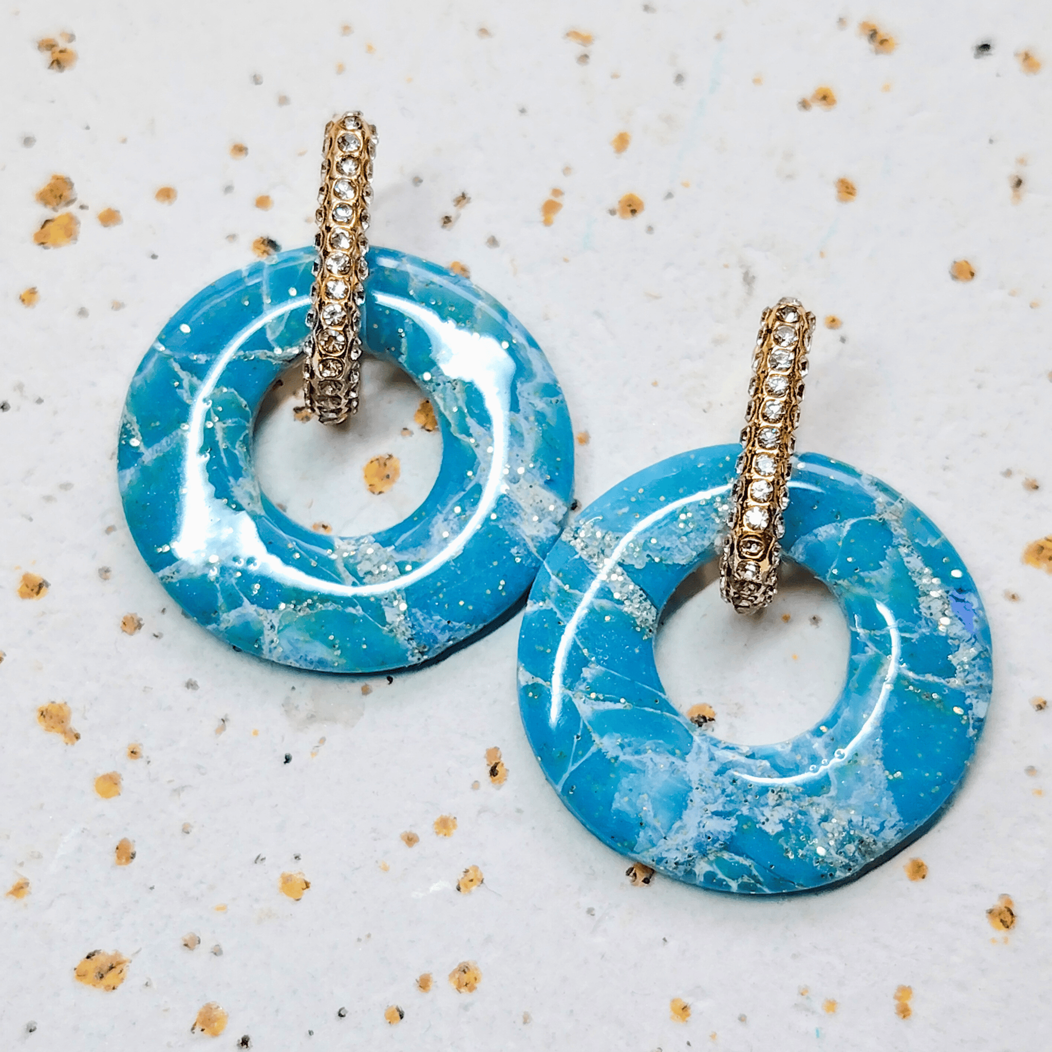 Paire de boucles d'oreilles créoles strass dorées avec leurs pampilles interchangeables Agate bleues et blanches aux reflets marbrés et pailletés d'or, posée sur un fond texturé.