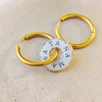 Deux boucles d'oreilles créoles dorées en acier inoxydable entrelacées avec une pampille blanche résinée ornée du mantra "SA TA NA MA" et de paillettes d'or.