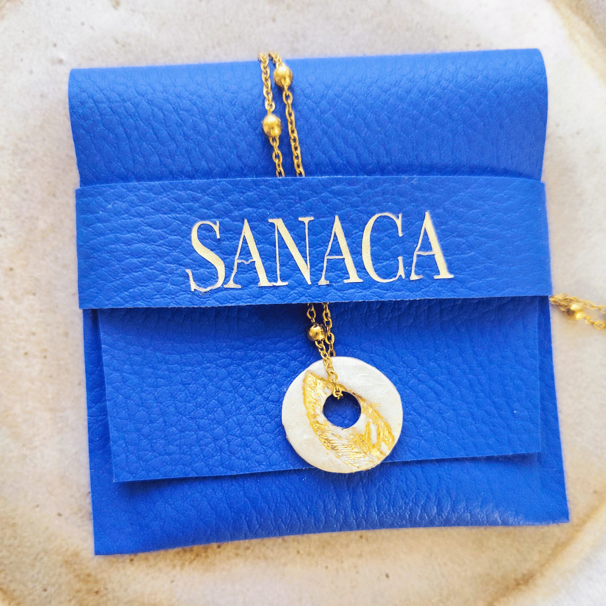 Pendentif Totem blanc et or posé sur la pochette signature en cuir bleu vif marquée du logo SANACA en lettres dorées, suspendu à sa chaîne en acier inoxydable doré.