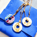 Trois pendentifs médaillons ronds Sanaca de styles différents, disposés sur une pochette signature en simili-cuir bleu électrique sous une lumière naturelle éclatante.