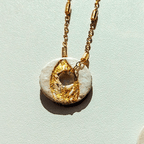 Vue rapprochée d'une pampille ronde artisanale en argile blanche, ornée d'un motif de feuille texturé à la feuille d'or, fixée à une fine chaîne dorée sur fond clair.