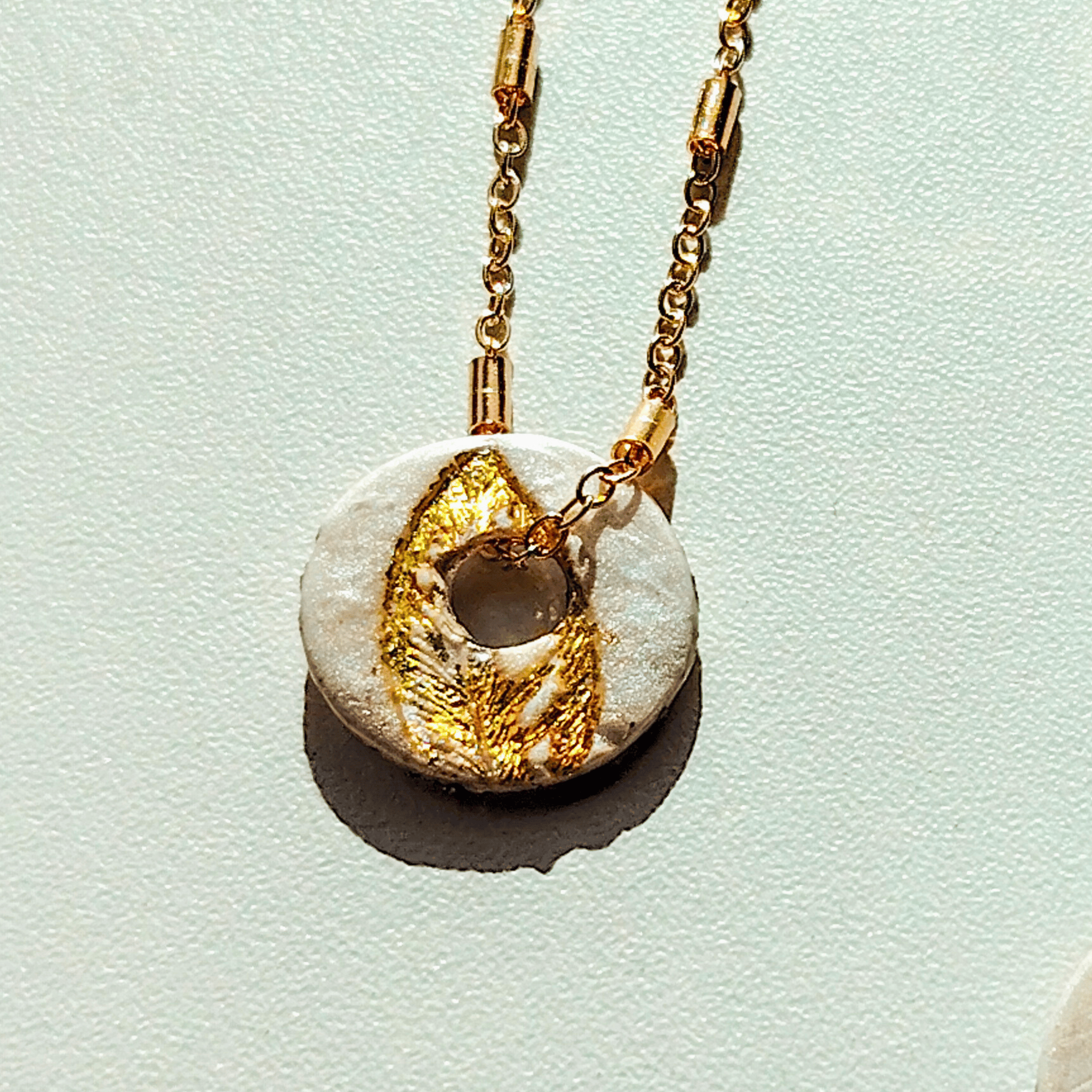 Vue rapprochée d'une pampille ronde artisanale en argile blanche, ornée d'un motif de feuille texturé à la feuille d'or, fixée à une fine chaîne dorée sur fond clair.