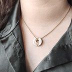 Gros plan d'un pendentif Totem rond et blanc texturé de feuille d'or, porté au cou sur une fine chaîne dorée par une femme portant une chemise vert kaki.
