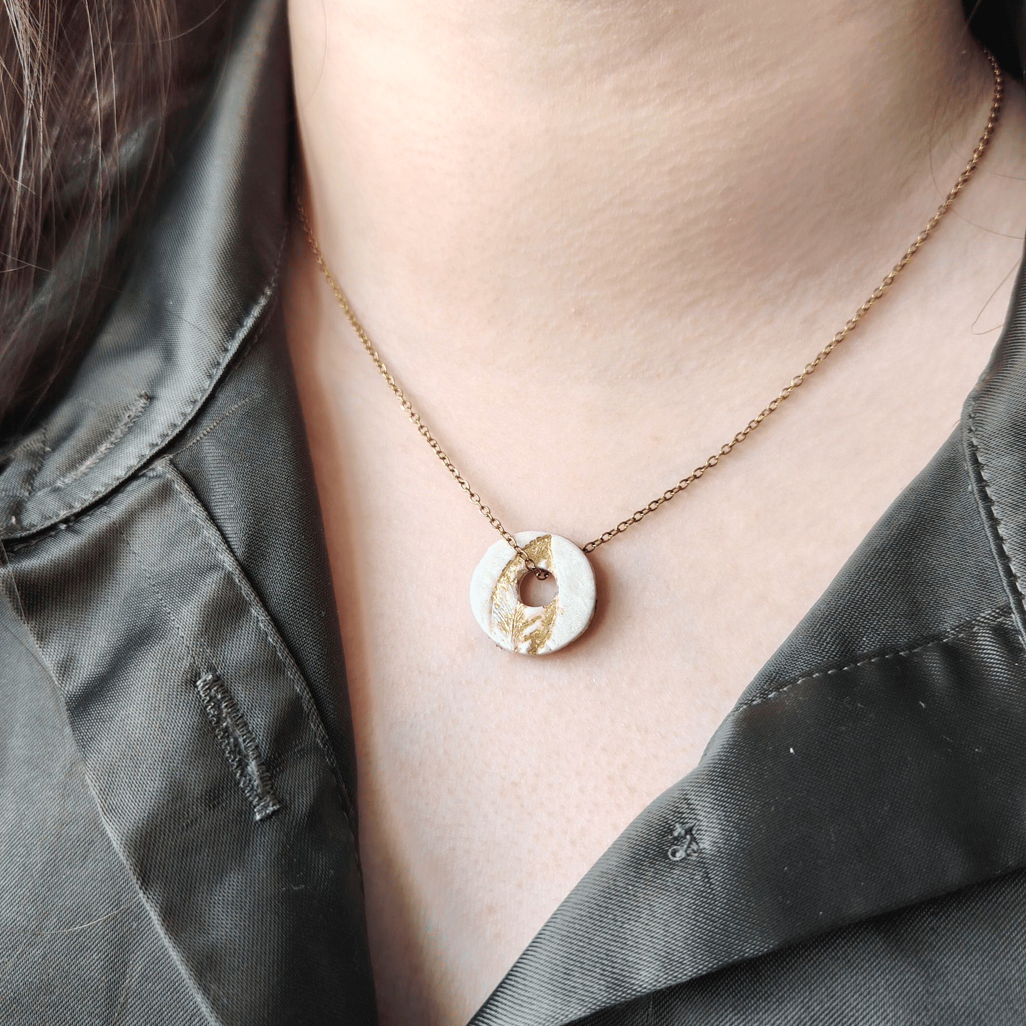 Gros plan d'un pendentif Totem rond et blanc texturé de feuille d'or, porté au cou sur une fine chaîne dorée par une femme portant une chemise vert kaki.