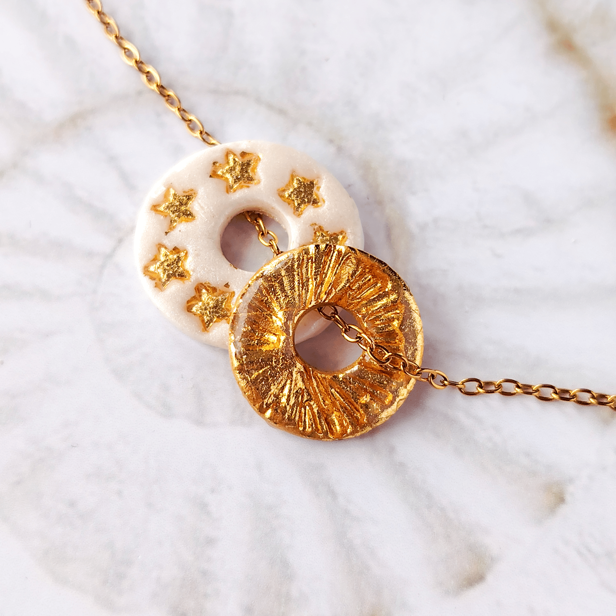 ccumulation de deux pendentifs totems artisanaux de la marque Sanaca sur une chaîne dorée : l'un en argile blanc nacré avec des étoiles dorées gravées, l'autre en finition entièrement texturée dorée effet émaillé.