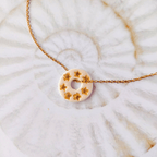 Pendentif totem rond en argile blanche à effet nacré orné de sept petites étoiles dorées, monté sur une fine chaîne en acier inoxydable doré, posé sur un fond texturé.