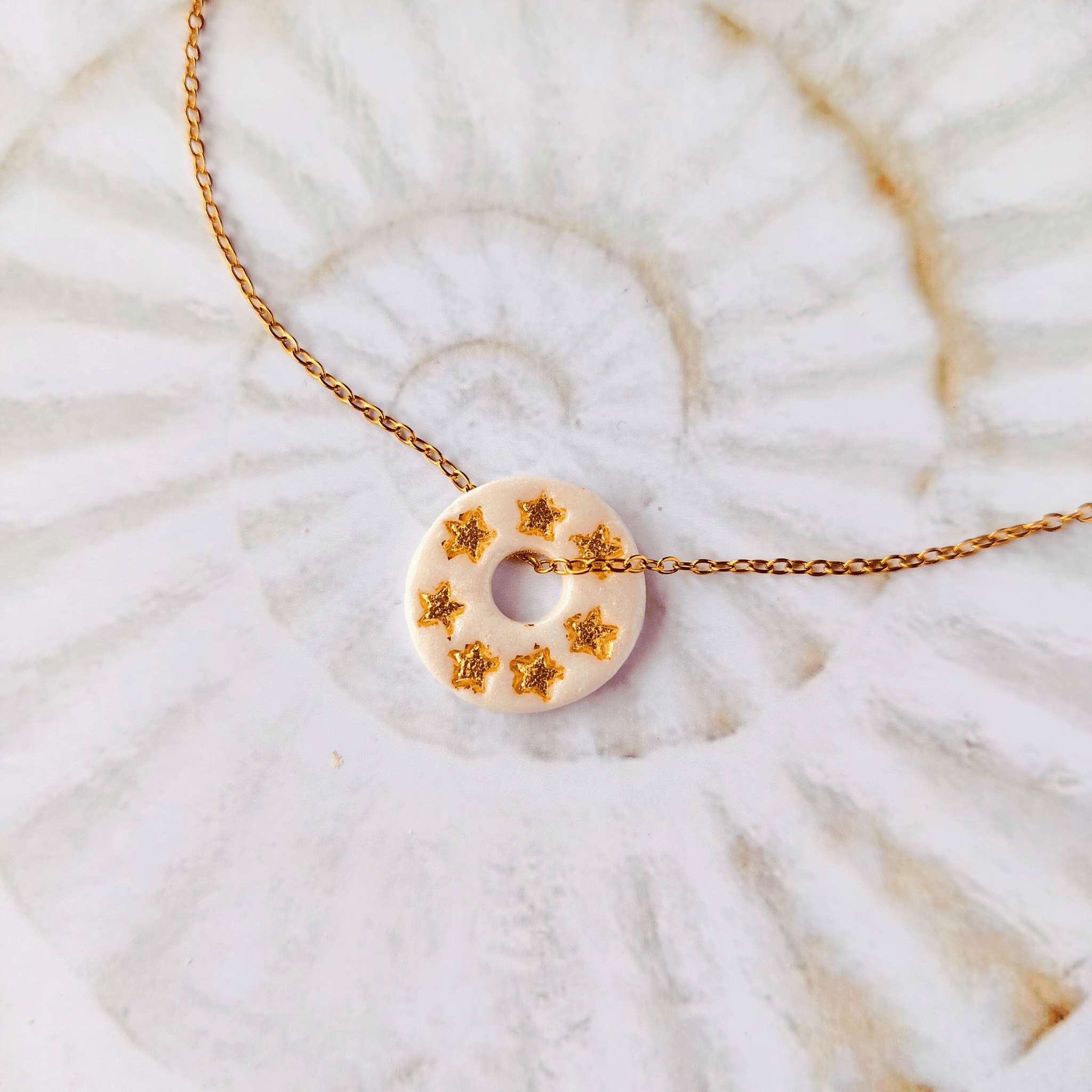 Pendentif totem rond en argile blanche à effet nacré orné de sept petites étoiles dorées, monté sur une fine chaîne en acier inoxydable doré, posé sur un fond texturé.