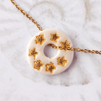 Gros plan sur le bijou artisanal fait main Totem Les Âmes, une pampille ronde de couleur blanche nacrée avec des motifs étoiles à la feuille d'or, glissée sur sa chaîne dorée à l'or fin.