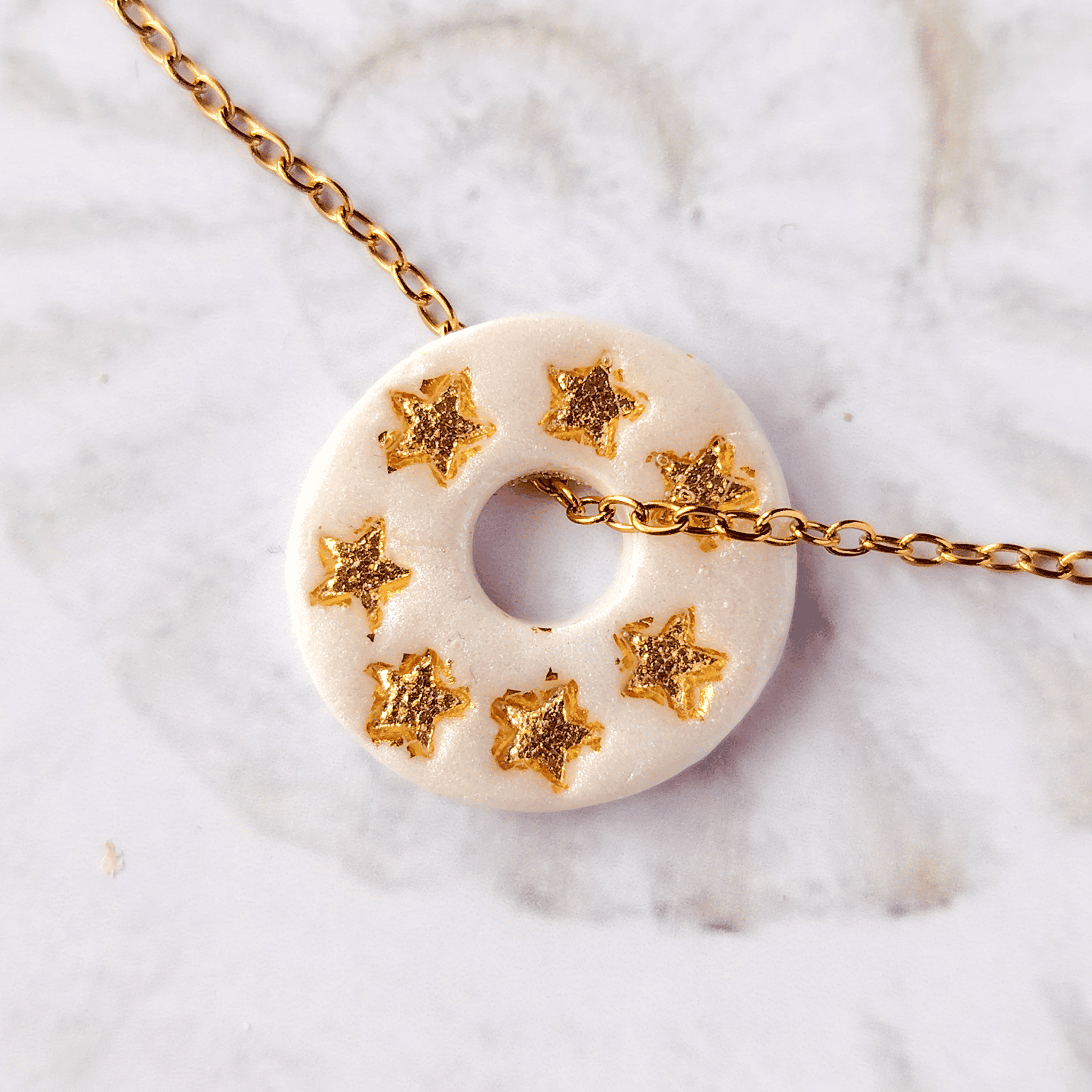 Gros plan sur le bijou artisanal fait main Totem Les Âmes, une pampille ronde de couleur blanche nacrée avec des motifs étoiles à la feuille d'or, glissée sur sa chaîne dorée à l'or fin.