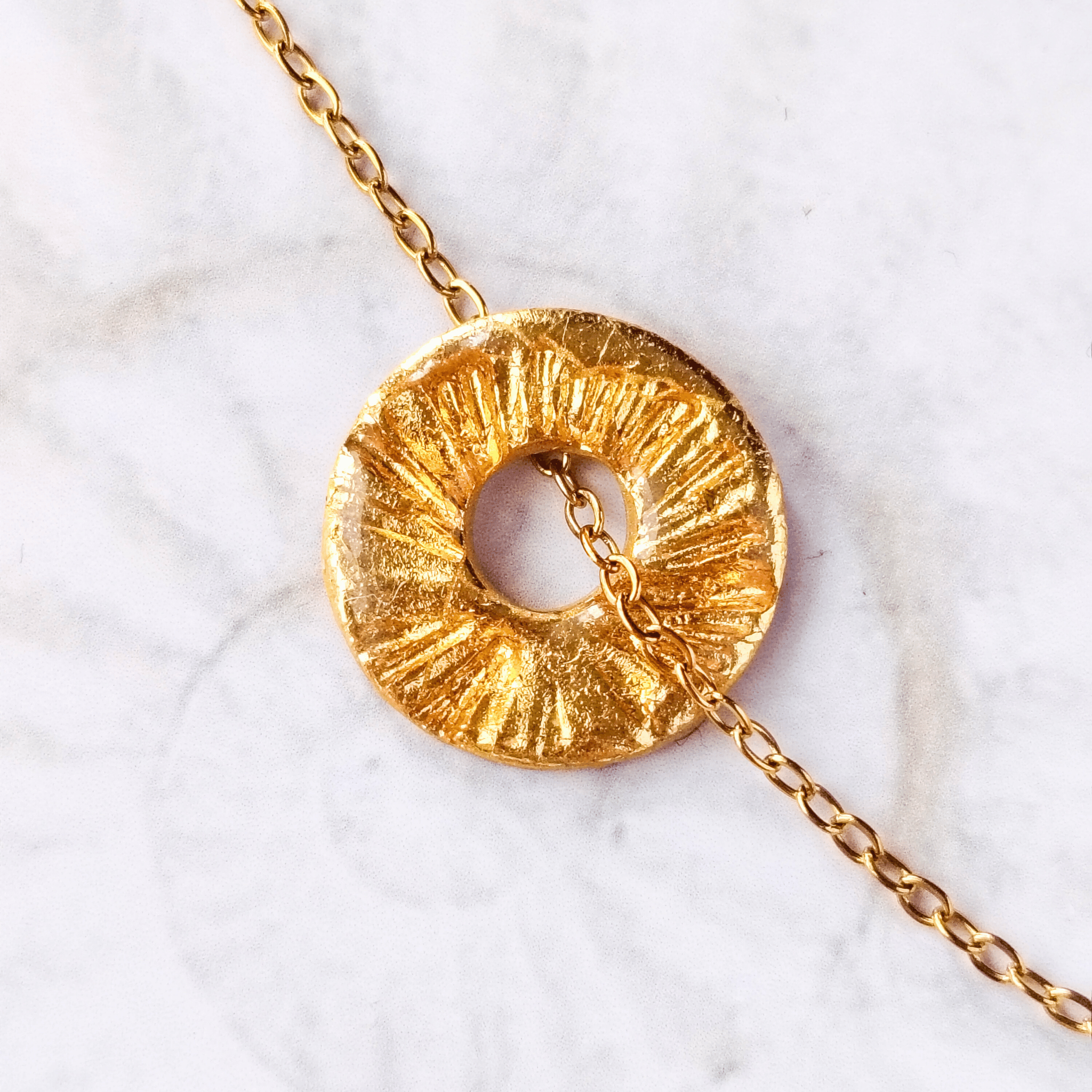Gros plan sur le pendentif Le Soleil SANACA, texture artisanale dorée à la feuille d'or et reflets de mica sur marbre blanc.