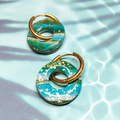 Boucles d'oreilles interchangeables Océane Flexilook en argile polymère et acier inoxydable, motif vagues bleu vert feuilles d'or, posées sur un fond aquatique clair.