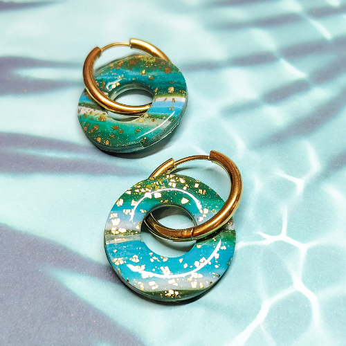 Boucles d'oreilles interchangeables Océane Flexilook en argile polymère et acier inoxydable, motif vagues bleu vert feuilles d'or, posées sur un fond aquatique clair.