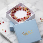 Une boîte cadeau en carton bleu clair, ornée du logo "Sanaca" en lettres dorées, est présentée sur un socle en marbre blanc. À l'intérieur, un collier 2 en 1 composé de perles rouges, orange et blanches est visible. Quelques perles rouges et dorées sont également éparpillées sur le marbre