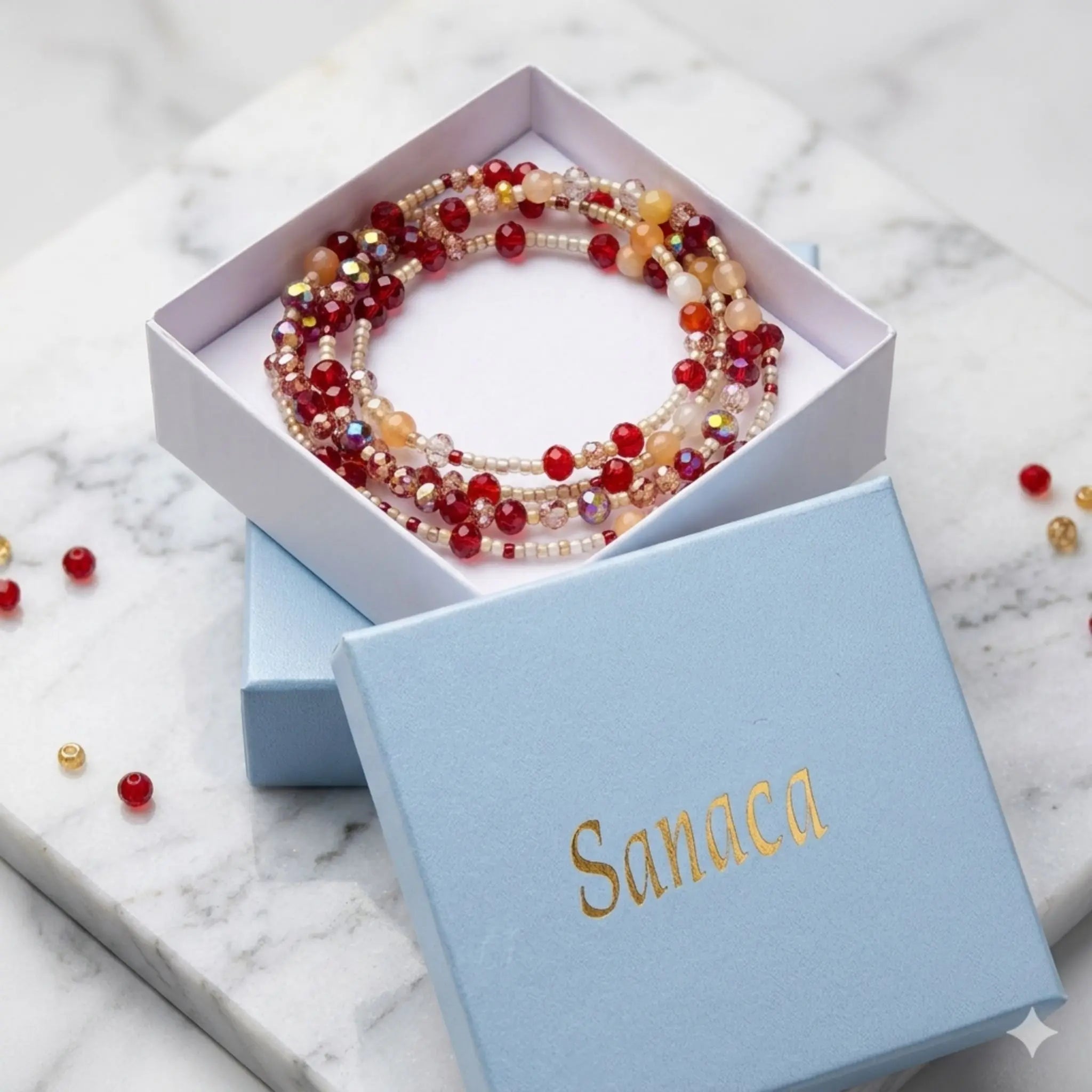 Une boîte cadeau en carton bleu clair, ornée du logo "Sanaca" en lettres dorées, est présentée sur un socle en marbre blanc. À l'intérieur, un collier 2 en 1 composé de perles rouges, orange et blanches est visible. Quelques perles rouges et dorées sont également éparpillées sur le marbre