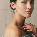 Portrait rapproché d'une femme à la peau claire portant la boucle d'oreille artisanale Sanaca en lapis-lazuli et aigue-marine. La photo met en valeur le port élégant du bijou et ses nuances bleues sur le lobe, avec une pose naturelle révélant l'épaule nue et un haut en lin à volants bleus. Fond texturé beige neutre