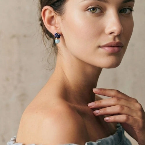 Portrait rapproché d'une femme à la peau claire portant la boucle d'oreille artisanale Sanaca en lapis-lazuli et aigue-marine. La photo met en valeur le port élégant du bijou et ses nuances bleues sur le lobe, avec une pose naturelle révélant l'épaule nue et un haut en lin à volants bleus. Fond texturé beige neutre