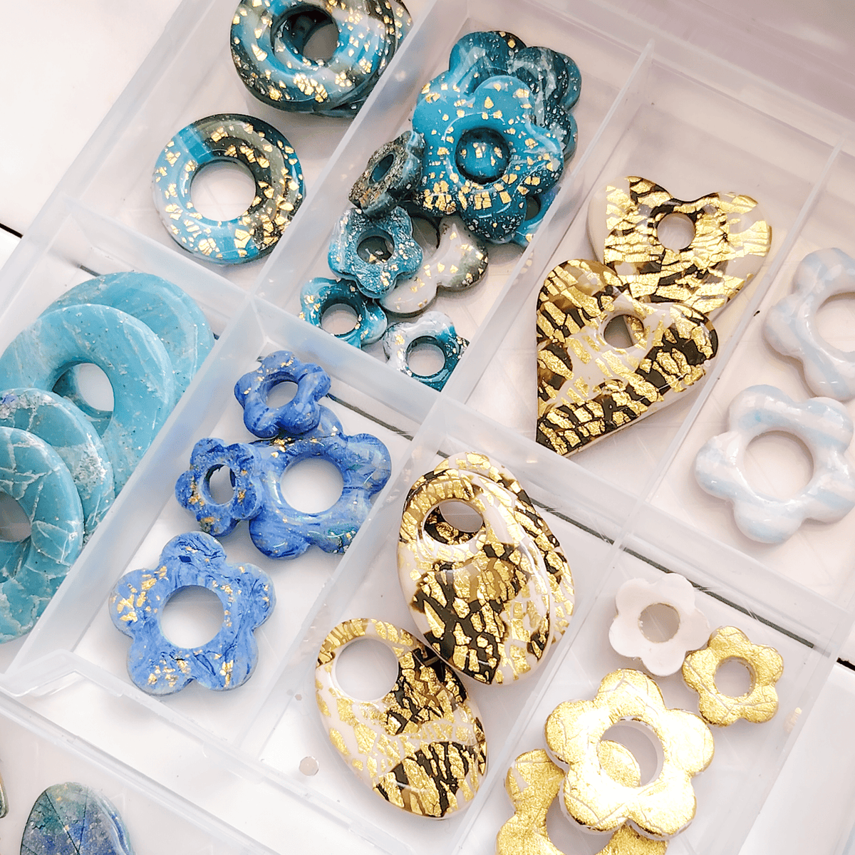 Gros plan détaillé de pampilles et pendentifs artisanaux Sanaca Bijoux rangés dans une boîte de compartiments transparente. La photo montre des breloques interchangeables pour le calendrier de l'Avent, avec des finitions marbrées bleues et des motifs dorés. Les pampilles en résine en forme de cœurs et de fleurs sont prêtes à être montées sur des créoles pour enfant.