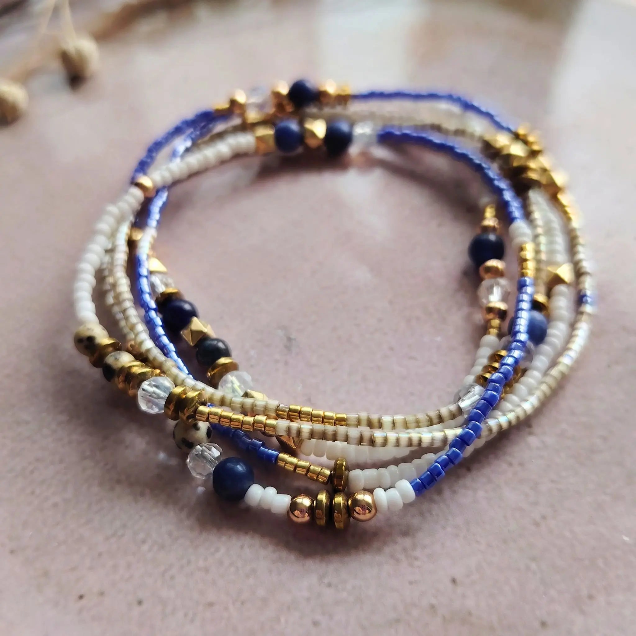 Un bracelet multi-tours pour femme enroulé, composé de perles naturelles de Lapis-Lazuli, de perles Miyuki blanches et d'hématite dorée, posé sur une surface texturée rose poudré.