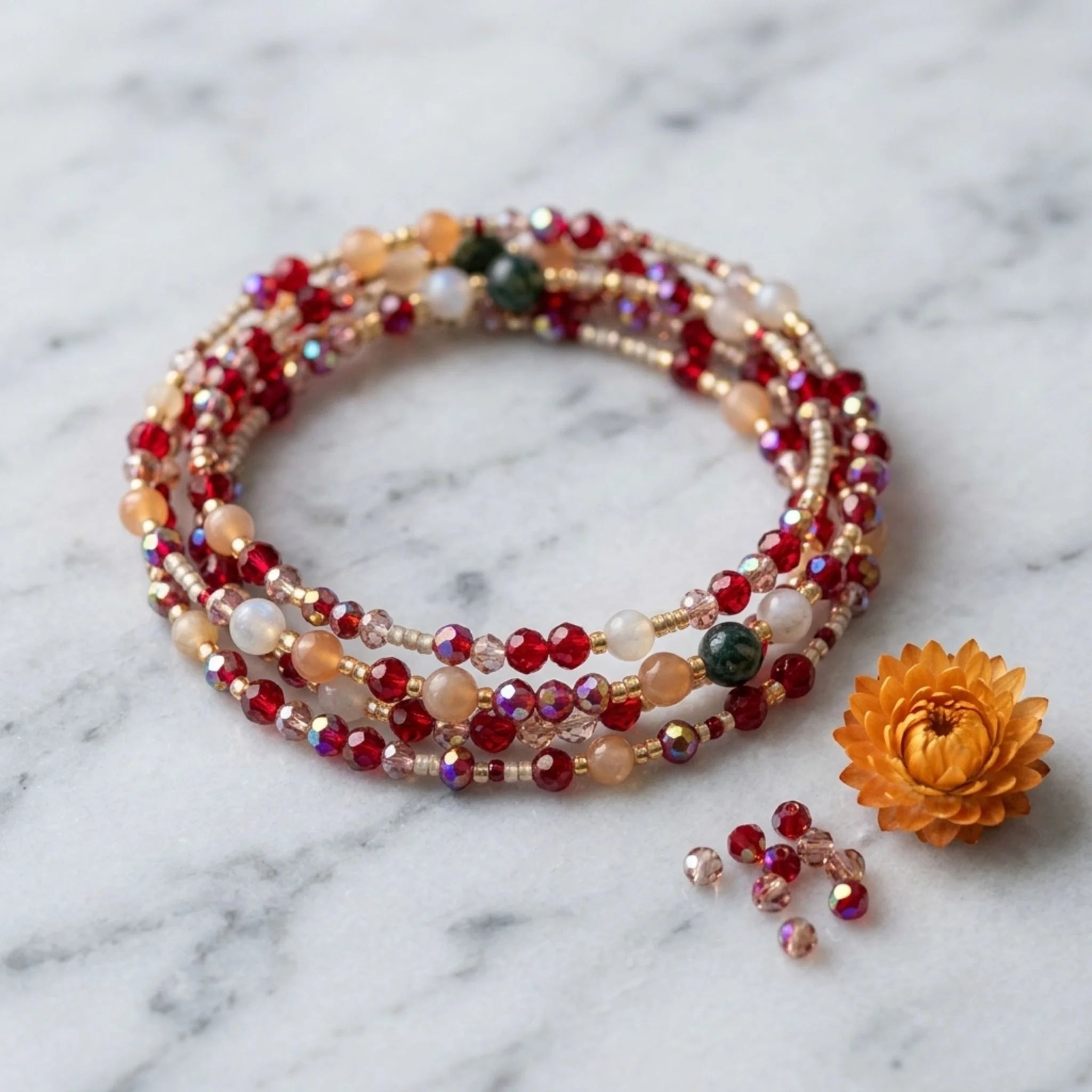 Le bracelet Charly de Sanaca Bijoux présenté en multi-tours sur une surface en marbre blanc brillant. Photographie haute définition de style magazine pro avec une fleur séchée orange, quelques perles rouges et dorées dispersées, et le logo doré Sanaca Bijoux.