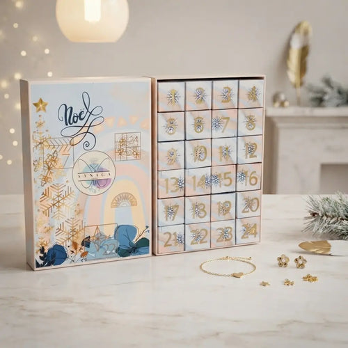 Calendrier de l'Avent bijoux Sanaca mis en scène sur un plan de travail en marbre avec des décorations de Noël. Le coffret est ouvert, révélant ses 24 cases numérotées[cite: 3, 5, 8]. Des bijoux dorés et pampilles interchangeables sont disposés devant, illustrant le concept créatif pour enfant. L'arrière-plan montre une ambiance chaleureuse avec une cheminée et des branches de sapin enneigées