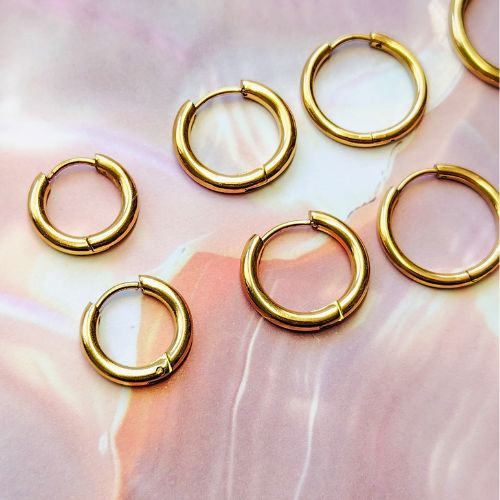 Gros plan sur les créoles Flexilook en acier inoxydable doré, présentées sur un fond nacré pour mettre en valeur leur éclat. Les modèles ronds et rectangulaires sont visibles, montrant la qualité et la finition des boucles d'oreilles.