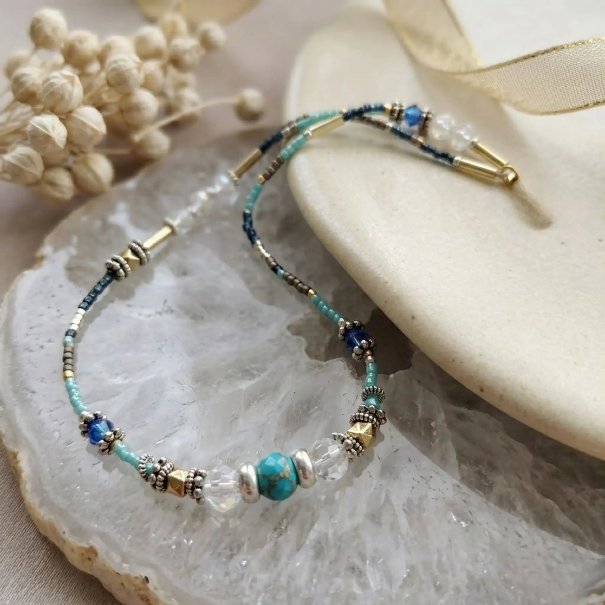 Le collier Luz repose sur une géode de cristal de quartz scintillant. Les détails des perles de turquoise, de cristal et d'or sont mis en valeur par la lumière naturelle. En arrière-plan, on voit une fleur séchée et un ruban doré sur une surface douce