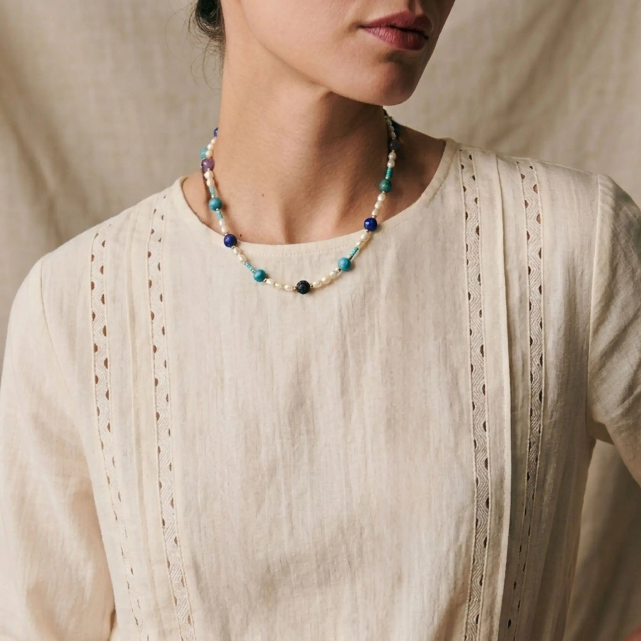 Collier Biarritz Sanaca porté au cou, mélange de pierres naturelles bleues et violettes avec perles d'eau douce sur une blouse en lin blanc.
