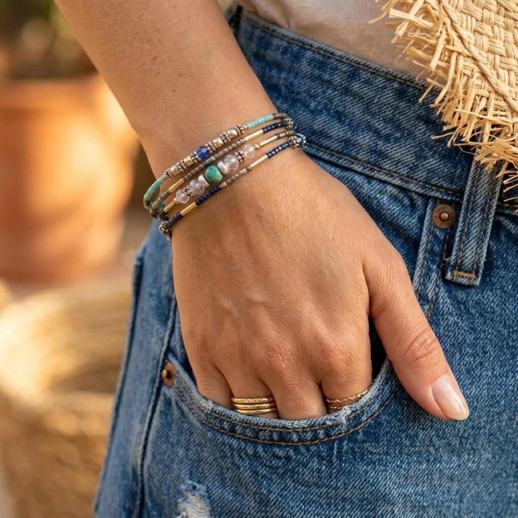 Une femme avec la main dans la poche de son jean en denim bleu porte le bracelet Luz au poignet. Le bracelet, avec ses perles turquoises, bleues et dorées, est enroulé plusieurs fois. On voit également d'autres bagues en or à ses doigts