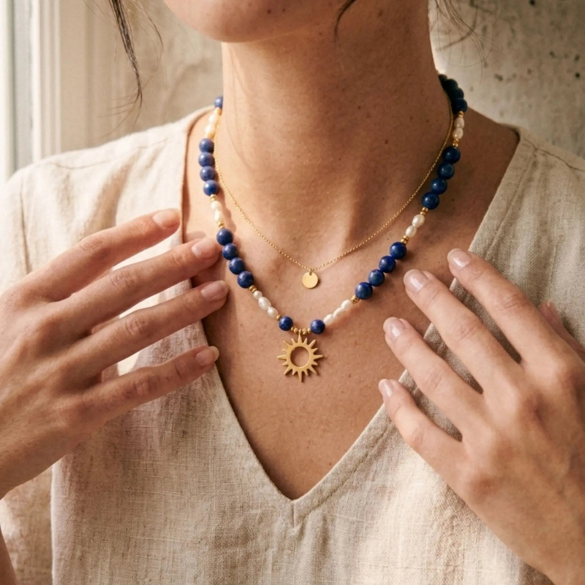 Portrait rapproché d'une femme portant le collier Solis en accumulation avec une fine chaîne dorée. Le contraste entre le bleu intense du lapis-lazuli, le blanc des perles d'eau douce et le lin de la blouse crée un look solaire et élégant typique du style artisanal du sud-ouest.