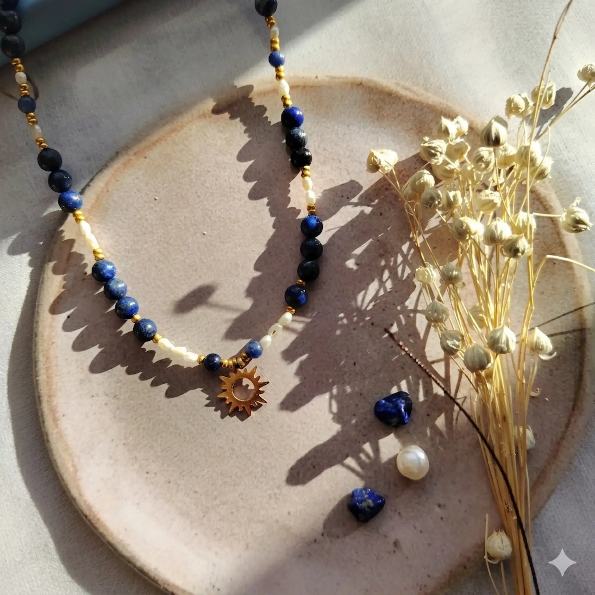 Collier artisanal Solis Sanaca composé de perles de lapis-lazuli bleu profond (6mm) et de perles d'eau douce nacrées. Le bijou est présenté sur une coupelle en céramique avec un pendentif soleil évidé en acier inoxydable doré, sous une lumière naturelle créant des ombres portées élégantes.