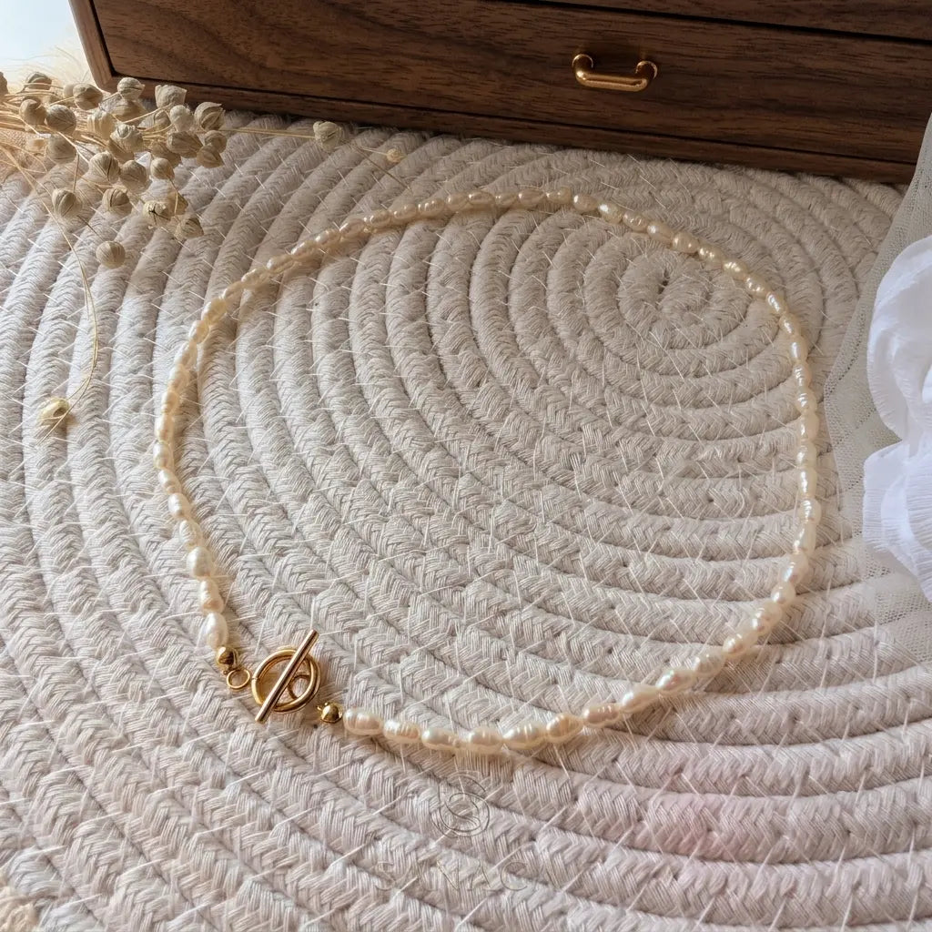 Vue de dessus d'un rang épuré de perles d'eau douce Sanaca formant un collier, reposant sur un plateau tressé rond et un pochon en lin, avec le fermoir T doré positionné sur le devant.