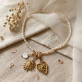 Collier en perles d'eau douce Sanaca disposé à plat sur un pochon en lin texturé avec logo SANACA estampé, accompagné de fleurs séchées, de nacre cœur et de pendentifs dorés soleil et cœur.