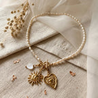 Collier en perles d'eau douce Sanaca disposé à plat sur un pochon en lin texturé avec logo SANACA estampé, accompagné de fleurs séchées, de nacre cœur et de pendentifs dorés soleil et cœur.
