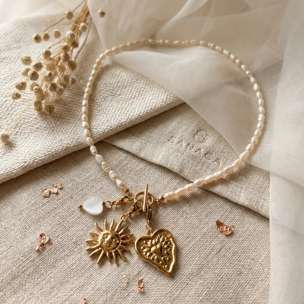 Collier en perles d'eau douce Sanaca disposé à plat sur un pochon en lin texturé avec logo SANACA estampé, accompagné de fleurs séchées, de nacre cœur et de pendentifs dorés soleil et cœur.