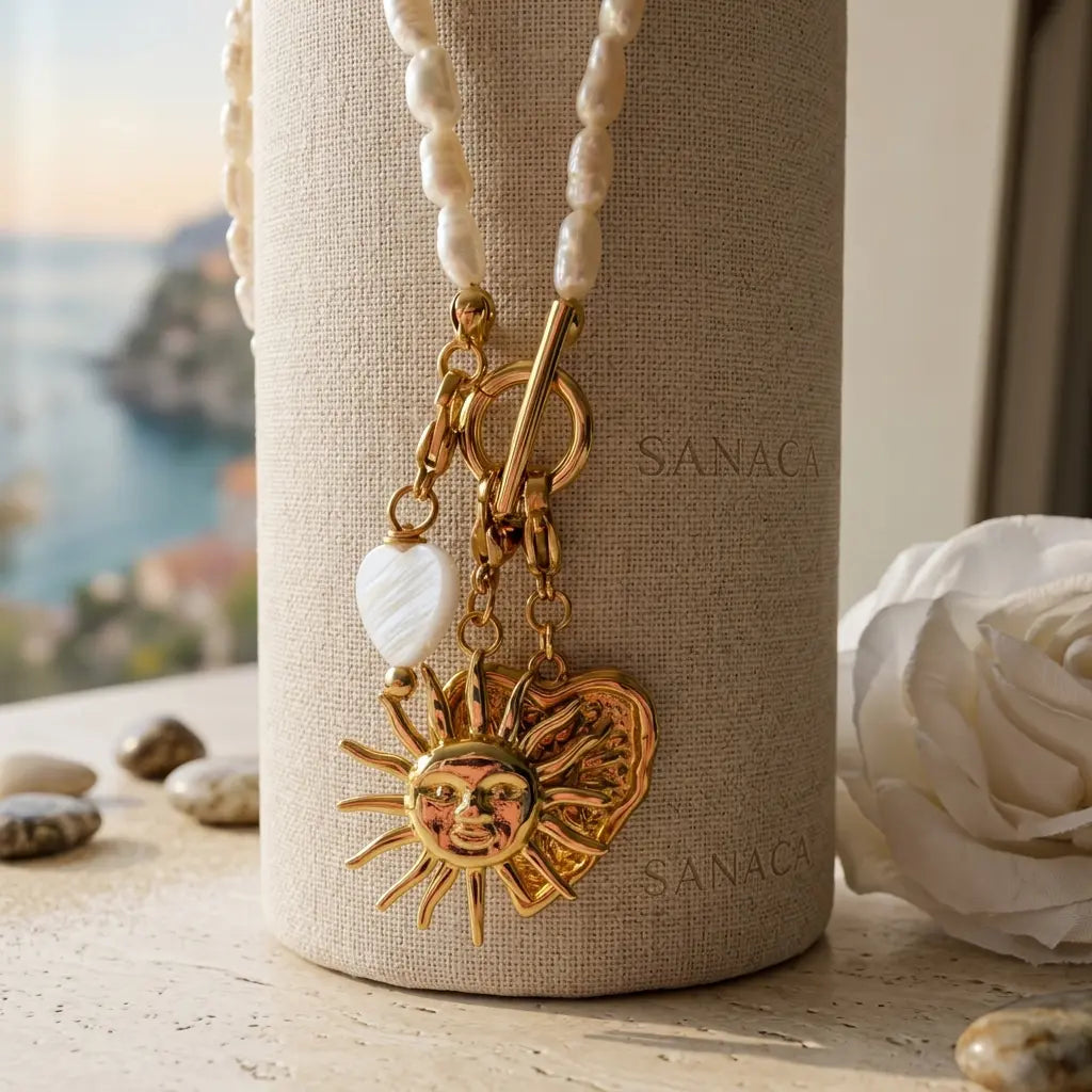 Détail du collier Sanaca avec fermoir T doré à l'or fin, orné d'un pendentif soleil sacralisé, d'un cœur gravé et d'un cœur en nacre blanche, devant un flacon texturé.