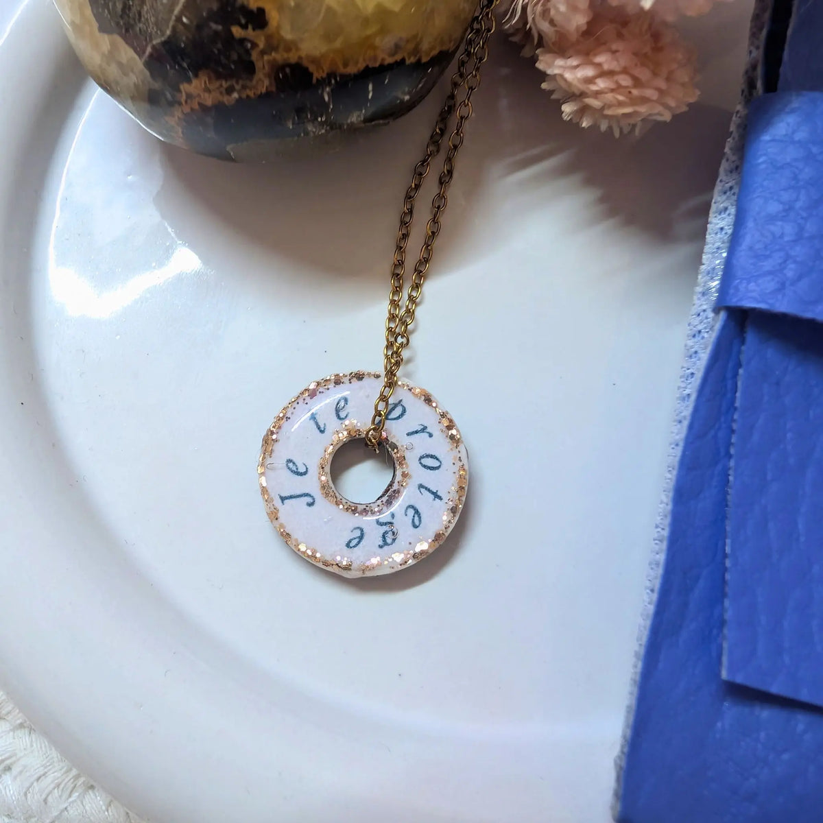 Vue rapprochée d'un collier artisanal. Un pendentif circulaire en résine blanche, bordé de paillettes dorées, est suspendu à une fine chaîne dorée. L'inscription manuscrite noire "Je te protège" est visible autour du trou central du pendentif. L'ensemble repose sur une surface blanche mate. En arrière-plan, flous, se trouvent une pierre polie, des fleurs séchées roses et une pochette en cuir bleu. La lumière est douce et naturelle, soulignant la texture des paillettes.