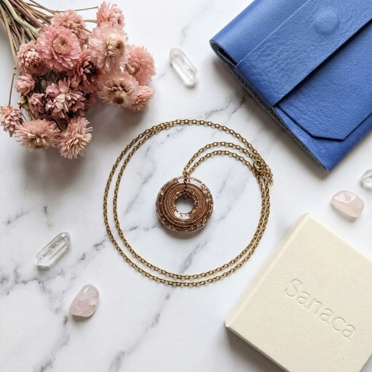 Composition photographique soignée vue de dessus, à plat, sur un plan de travail en marbre blanc. Un collier avec un pendentif circulaire "beignet" en résine marron-bronze pailleté est centré, sa fine chaîne dorée formant une boucle élégante autour de lui. Autour, sont disposés harmonieusement : un bouquet de fleurs séchées roses, plusieurs cristaux de quartz bruts, une pochette en cuir bleu vif et une boîte-cadeau kraft beige estampillée discrètement "Sanaca". L'ambiance est artisanale, naturelle et chic.