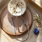 Set de bijoux artisanaux comprenant le collier sautoir et le bracelet Marie assorti en Lapis-Lazuli et doré, présentés dans une coupelle floutée sur un plateau en terre cuite avec une pierre brute de lapis-lazuli.