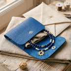 Présentation du collier Solis délicatement déposé dans sa pochette de protection en simili cuir bleu roi avec logo Sanaca doré. L'image inclut des éléments de l'atelier comme une enveloppe en lin et des bobines de fil, soulignant le caractère "Pièce Unique" et prêt-à-offrir.