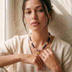 Portrait d'un mannequin portant le collier solaire Solis de Sanaca. L'image met en valeur l'alternance des pierres bleues de lapis-lazuli et des perles d'eau douce blanches sur une blouse en lin beige, illustrant le tombé parfait du pendentif soleil doré sur le décolleté