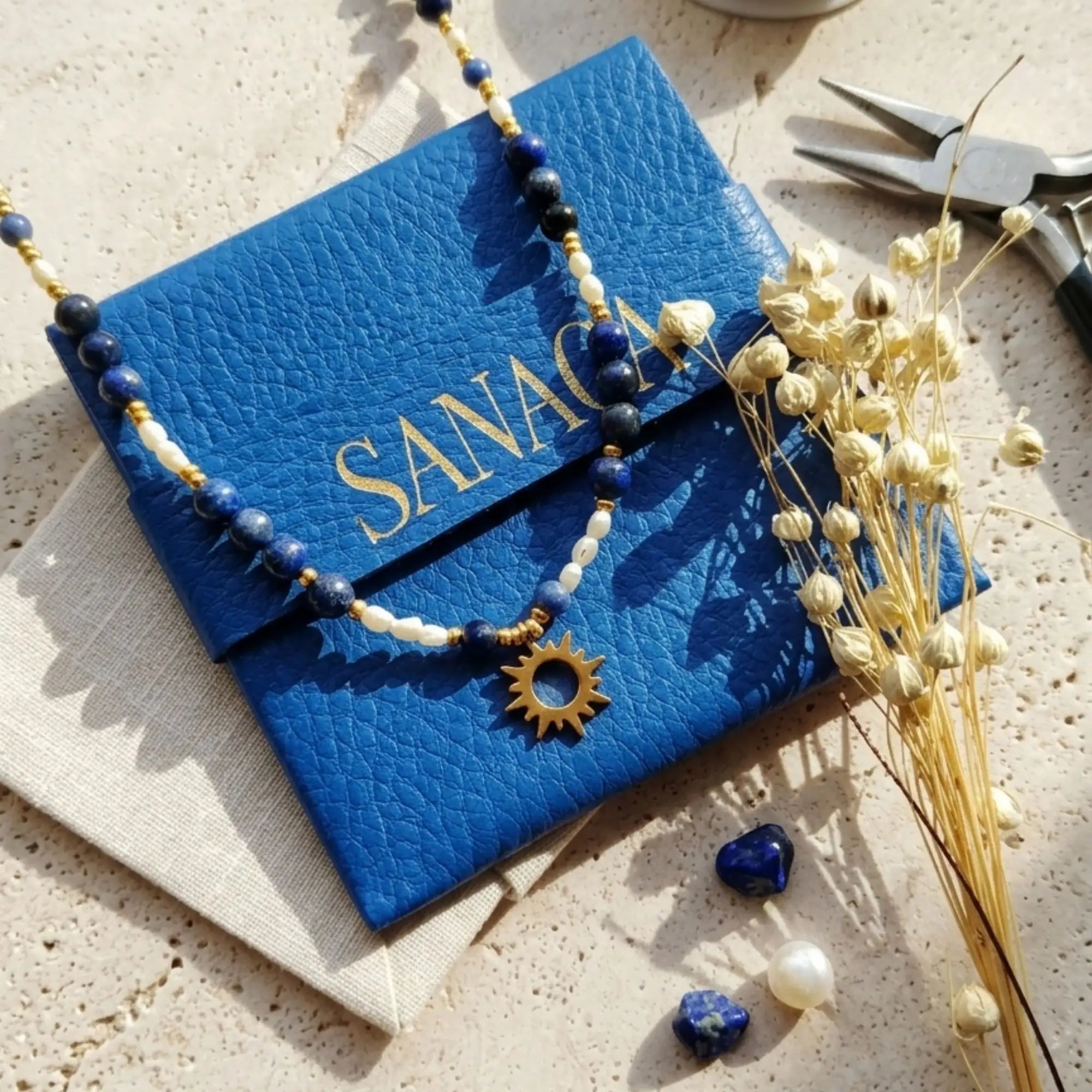Vue d'atelier du collier Solis Sanaca : perles de lapis-lazuli et perles d'eau douce sur une pochette en cuir bleu signature. La scène, baignée de lumière naturelle, inclut des outils de bijoutier et des fleurs séchées, soulignant une fabrication artisanale dans le sud-ouest.
