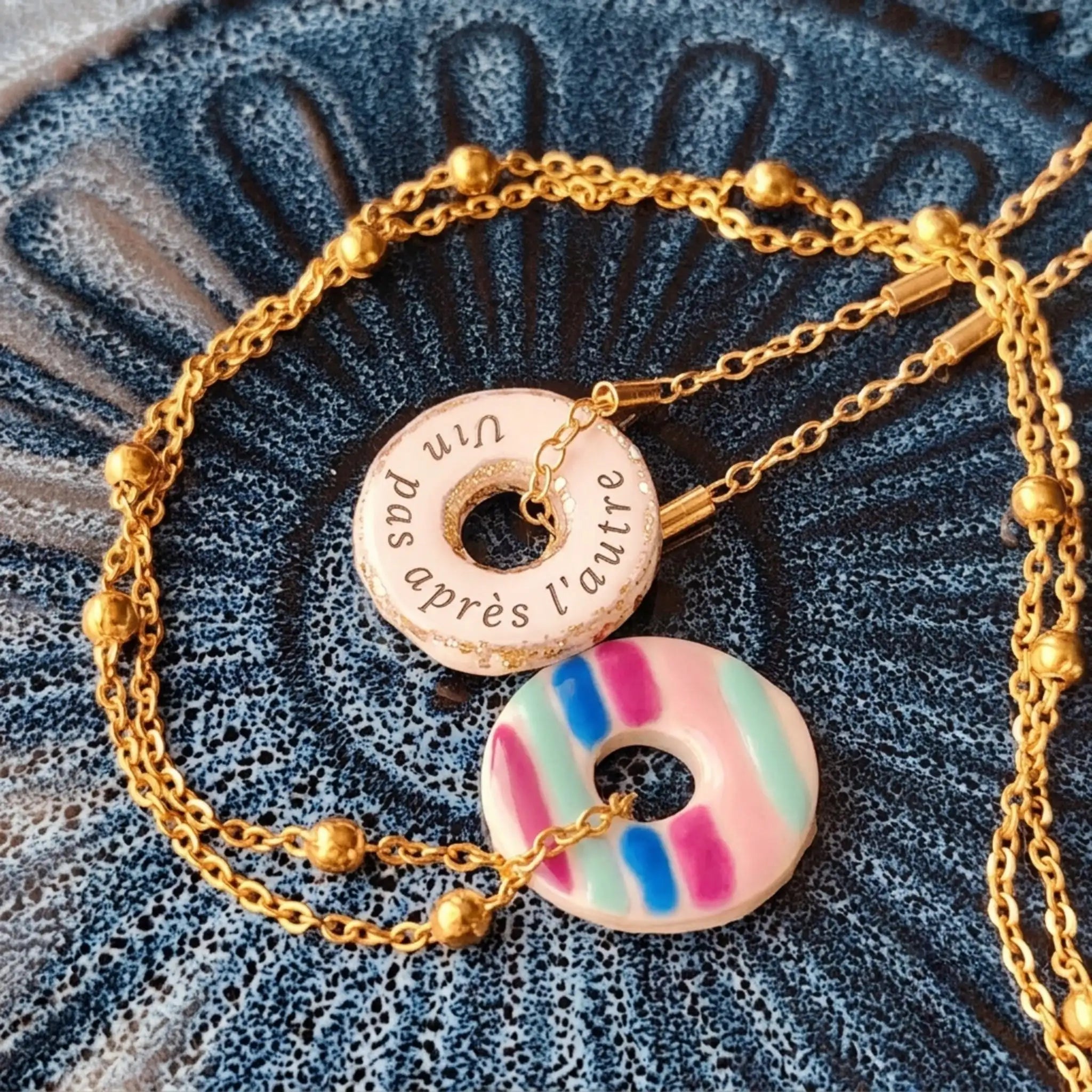 Une photographie en gros plan, prise en plongée, montre deux colliers à pendentif en forme de beignet reposant sur une surface en céramique texturée bleu foncé. Les deux colliers sont dotés d'une chaîne en or avec de petites perles dorées. Le pendentif du haut est un beignet en céramique blanche avec un centre creux et un saupoudrage de paillettes dorées sur le bord extérieur. Une phrase en français est imprimée en lettres noires autour du centre : "Un pas après l'autre". Le pendentif du bas est un beignet 