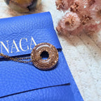 Gros plan détaillé d'un pendentif circulaire en résine marron-bronze pailleté reposant sur une pochette en cuir bleu vif à la texture grainée. La fine chaîne dorée du collier est partiellement visible, passant dans le trou du pendentif. Une partie du nom de la marque "NACA" (de "Sanaca") est estampillée en lettres dorées sur la pochette, juste au-dessus du pendentif. En arrière-plan, légèrement floues, se trouvent des fleurs séchées roses. L'éclairage direct fait scintiller les paillettes du pendentif.